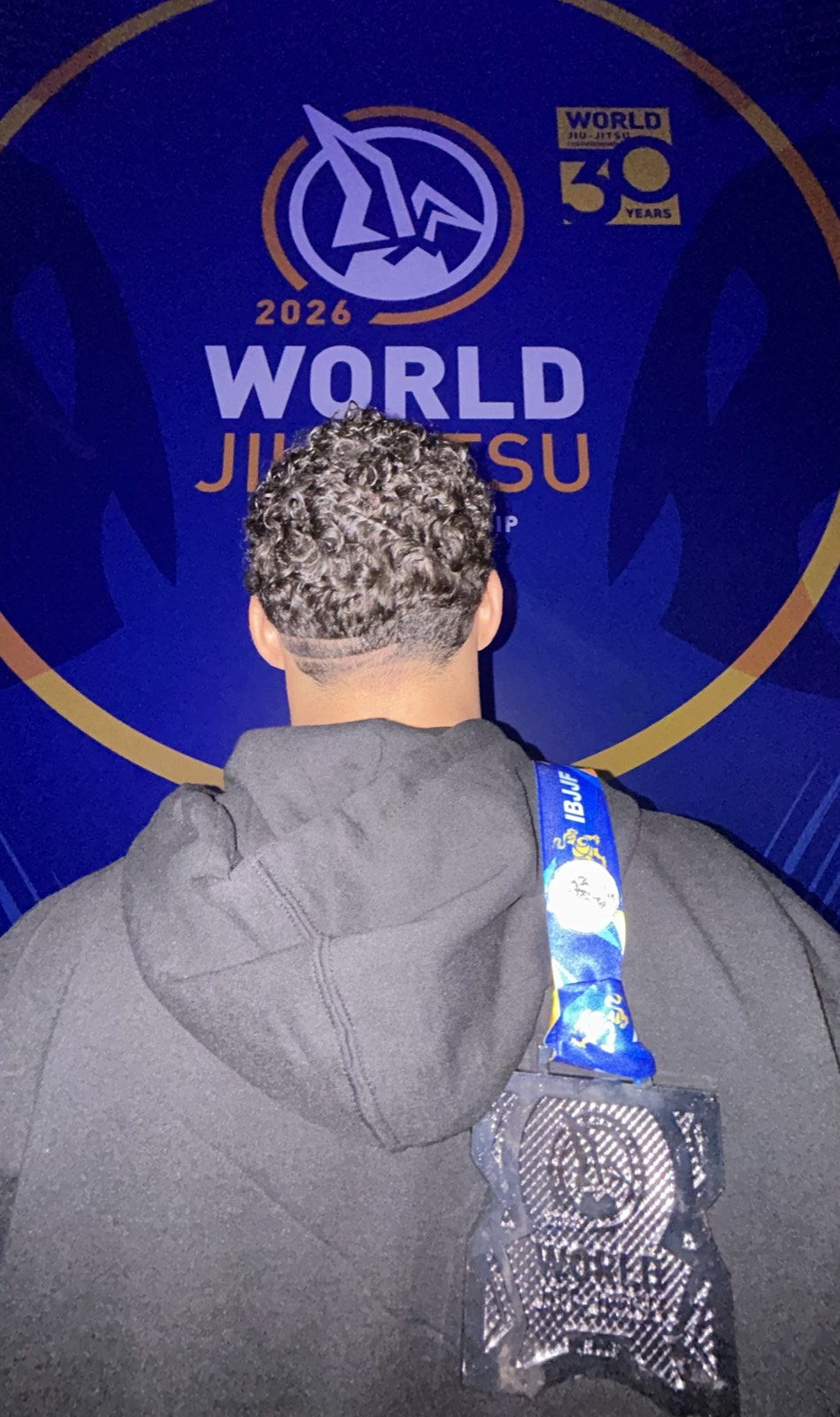 Medalha do Mundial IBJJF 2026 pendurada nas costas de Antonio diante do painel do World Jiu-Jitsu